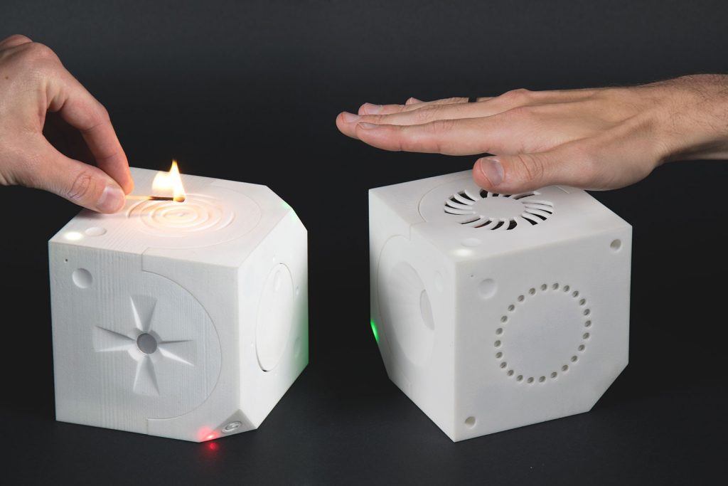 Loaded Dice, left is the sensor cube with a temperature sensor facing upwards, sensing heat from a match, actuator cube on the right with a ventilator actuator facing up and a hand feeling that air